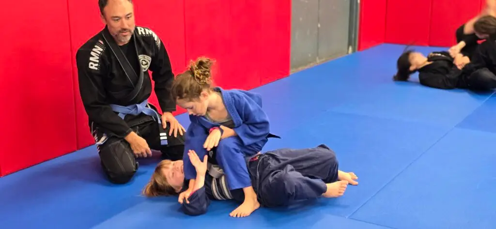 Brazilian Jiu-Jitsu coach guiding two children practicing a grappling technique at Frequency Martial Arts in Bloomington, Illinois.
